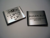 Zwei Metallplatten, eine mit einem Auge und Braille-Punkten, die andere mit der Aufschrift AUGEN AUF.