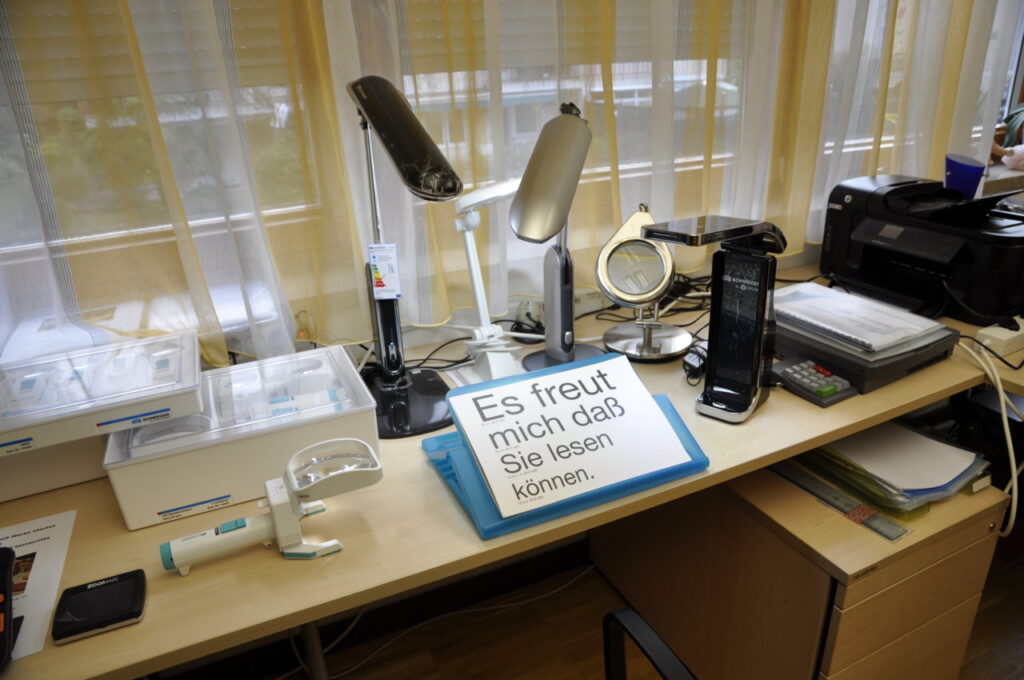 Ein Schreibtisch voller nützlicher Hilfsmittel für Menschen mit Seheinschränkungen, darunter Lupen und Leuchten, mit einem großen Schild: Es freut mich daß Sie lesen können.