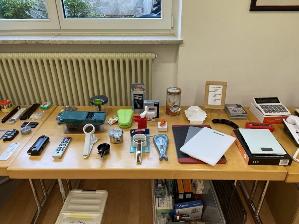 Ein Tisch mit verschiedenen Hilfsmitteln zur Unterstützung im Alltag, darunter Küchenutensilien, ein Taschenrechner, und beschriftete Behälter, steht an einem Fenster.