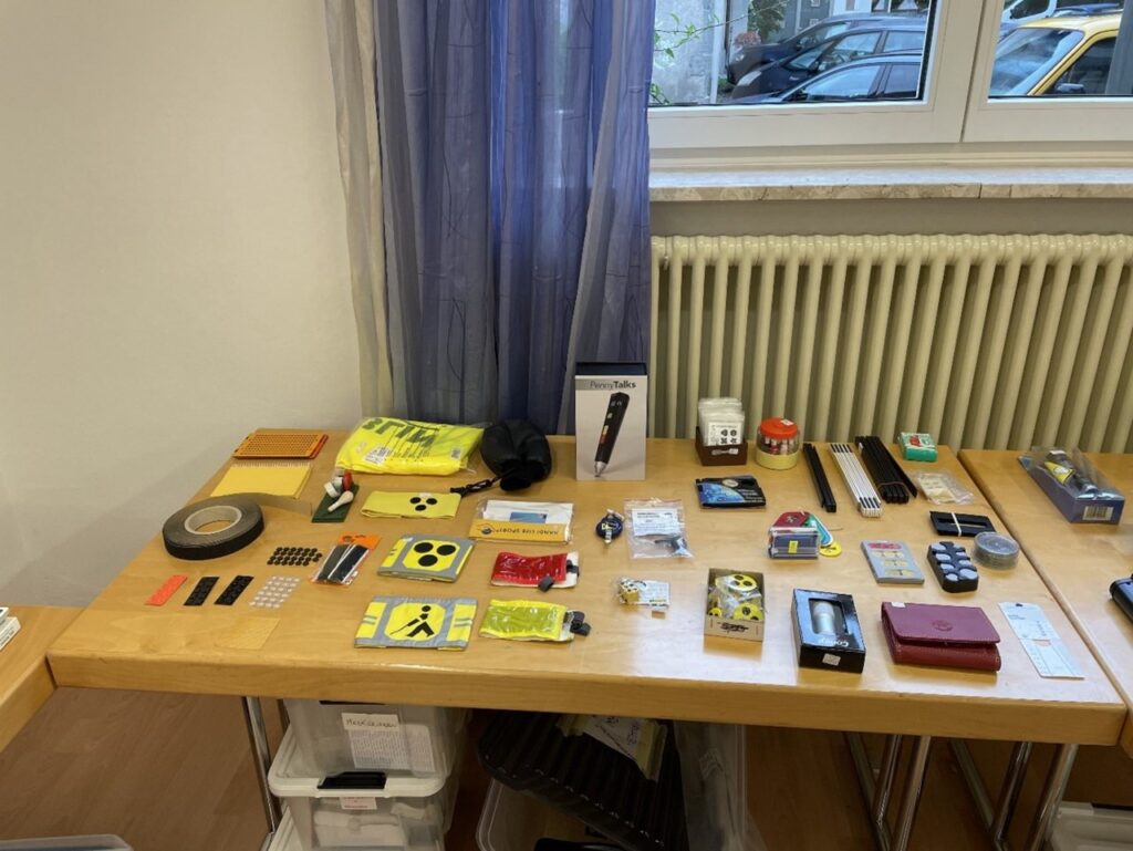 Auf einem Tisch liegen verschiedene Hilfsmittel für Menschen mit Sehbehinderung, wie taktile Markierungen und ein elektronisches Lesegerät, ordentlich nebeneinander.