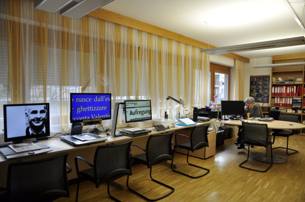 Ein Büro mit mehreren Computern, auf deren Bildschirmen vergrößerter Text angezeigt wird, und einer Person, die telefoniert.