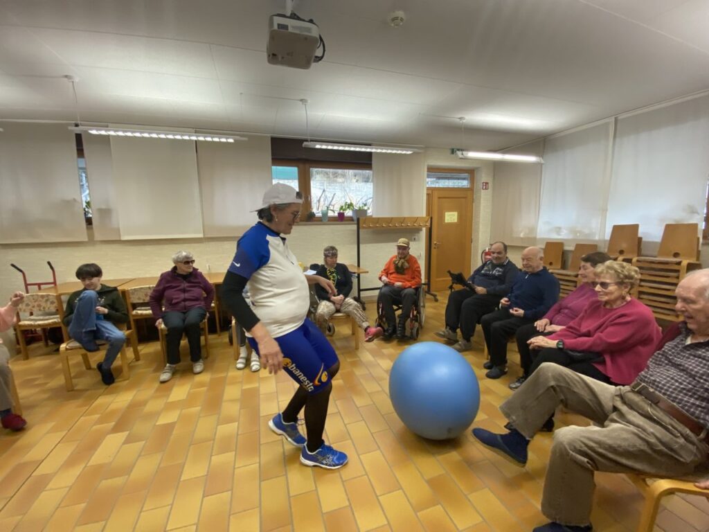 Eine Gruppe von Senioren sitzt im Kreis und nimmt an einer Bewegungsstunde mit einem großen blauen Gymnastikball teil.