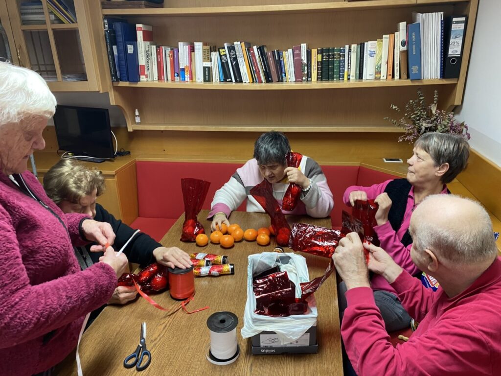 Eine Gruppe älterer Menschen sitzt um einen Tisch und verpackt Geschenke und Mandarinen in rotes Papier in einem Raum mit Bücherregal.
