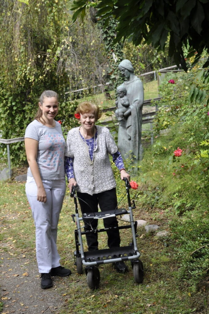Eine ältere Frau mit Rollator steht neben einer jüngeren Frau im Garten vor einer Statue.