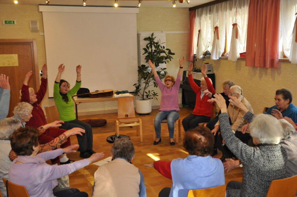 Eine Gruppe älterer Menschen sitzt in einem Kreis und macht gemeinsame Gymnastikübungen mit erhobenen Armen in einem hellen Raum.