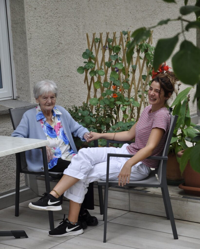 Eine ältere Frau und eine jüngere Frau sitzen draußen auf Stühlen, halten sich an den Händen und lächeln.