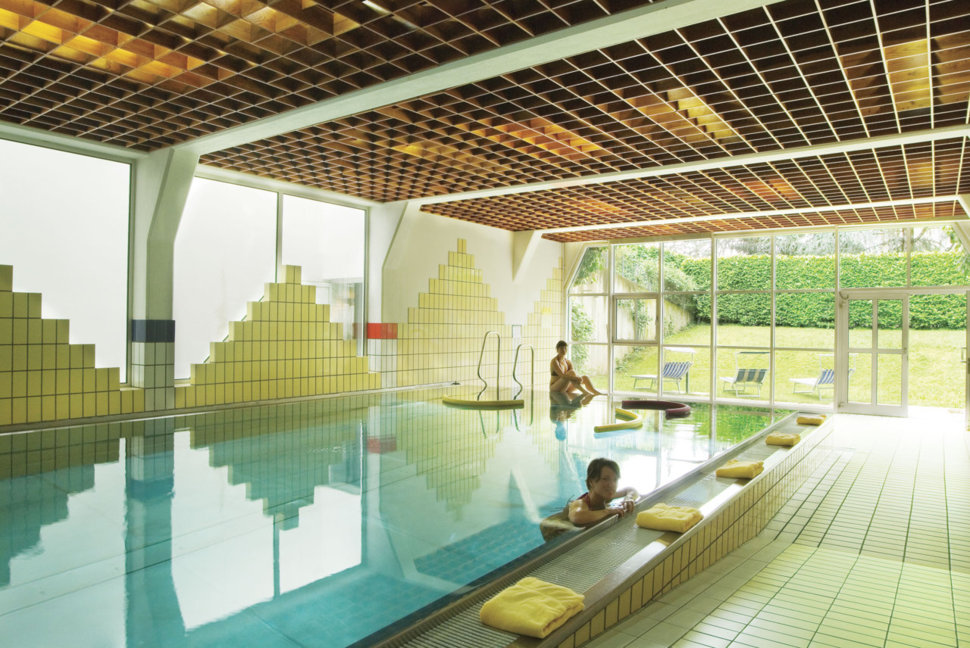 Ein Hallenbad mit gelben Fliesen, einer Glasfront und mehreren entspannten Menschen im und am Wasser.