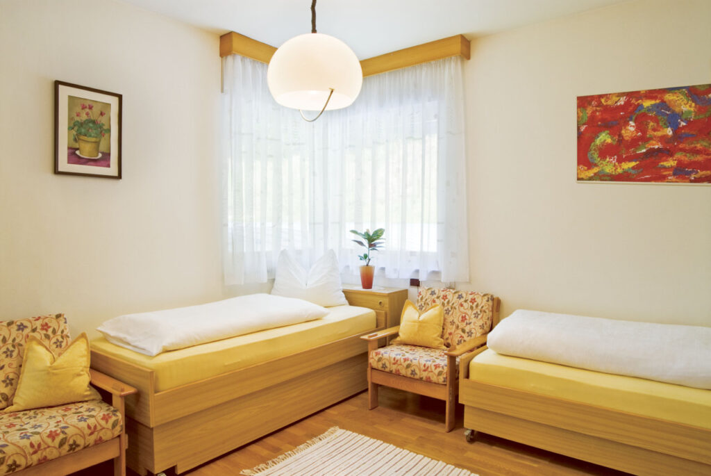 Ein helles Zimmer mit zwei Einzelbetten, zwei Sesseln und einem kleinen Nachttisch mit einer Pflanze, dekoriert mit Bildern an den Wänden.