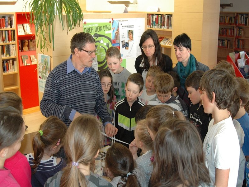 Ein Mann erklärt einer Gruppe von Kindern in einer Bibliothek etwas an einem Tisch.