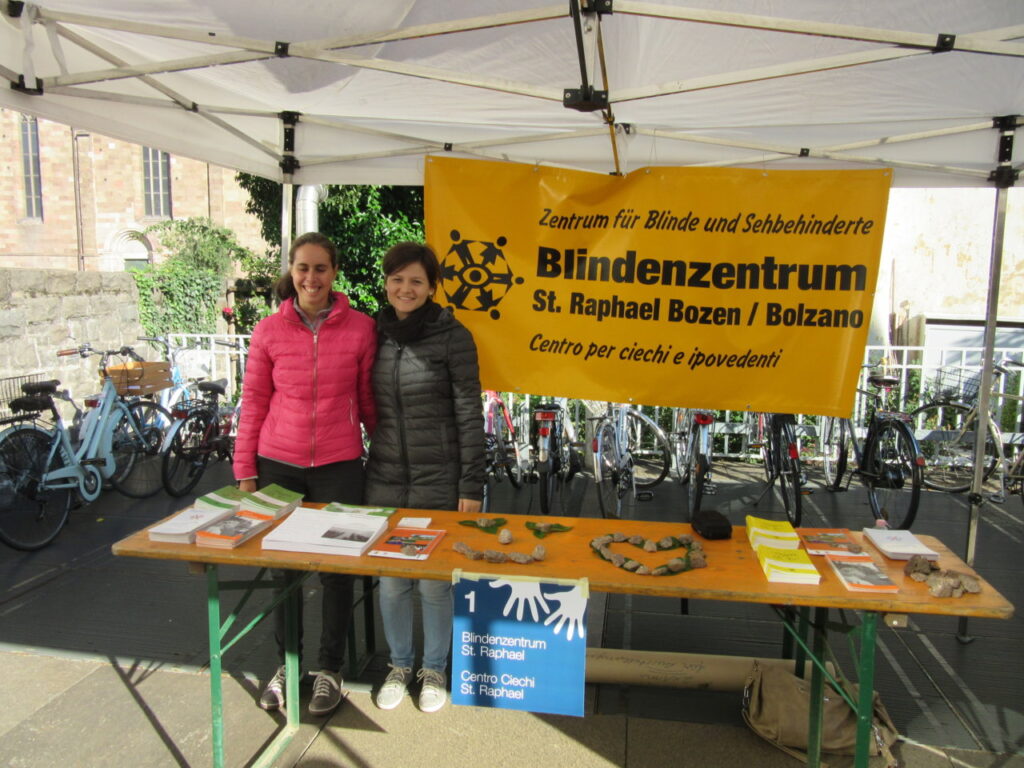 Zwei Personen stehen an einem Infostand des Blindenzentrums St. Raphael Bozen unter einem weißen Pavillon, der von Fahrrädern umgeben ist.