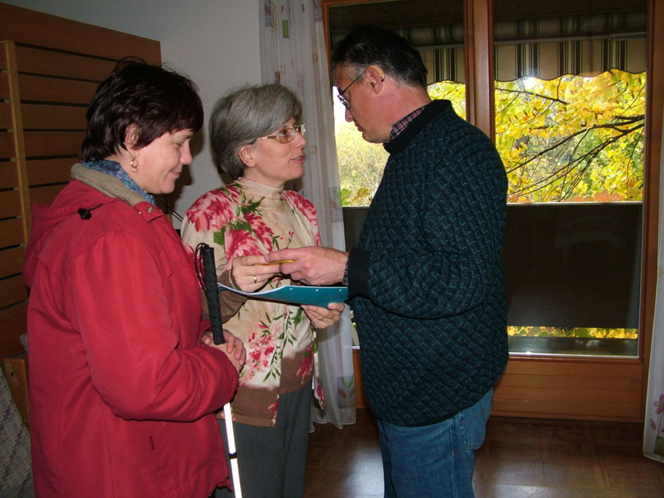 Eine Frau mit rotem Mantel und Blindenstock spricht mit einer Frau in geblümter Bluse und einem Mann in dunkelblauem Pullover, während sie gemeinsam ein Dokument betrachten.