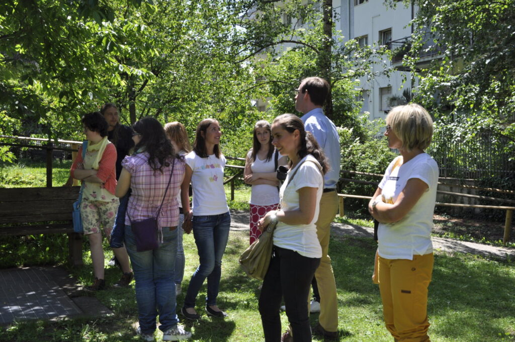 Eine Gruppe von Menschen steht bei sonnigem Wetter in einem grünen Garten zusammen und unterhält sich.