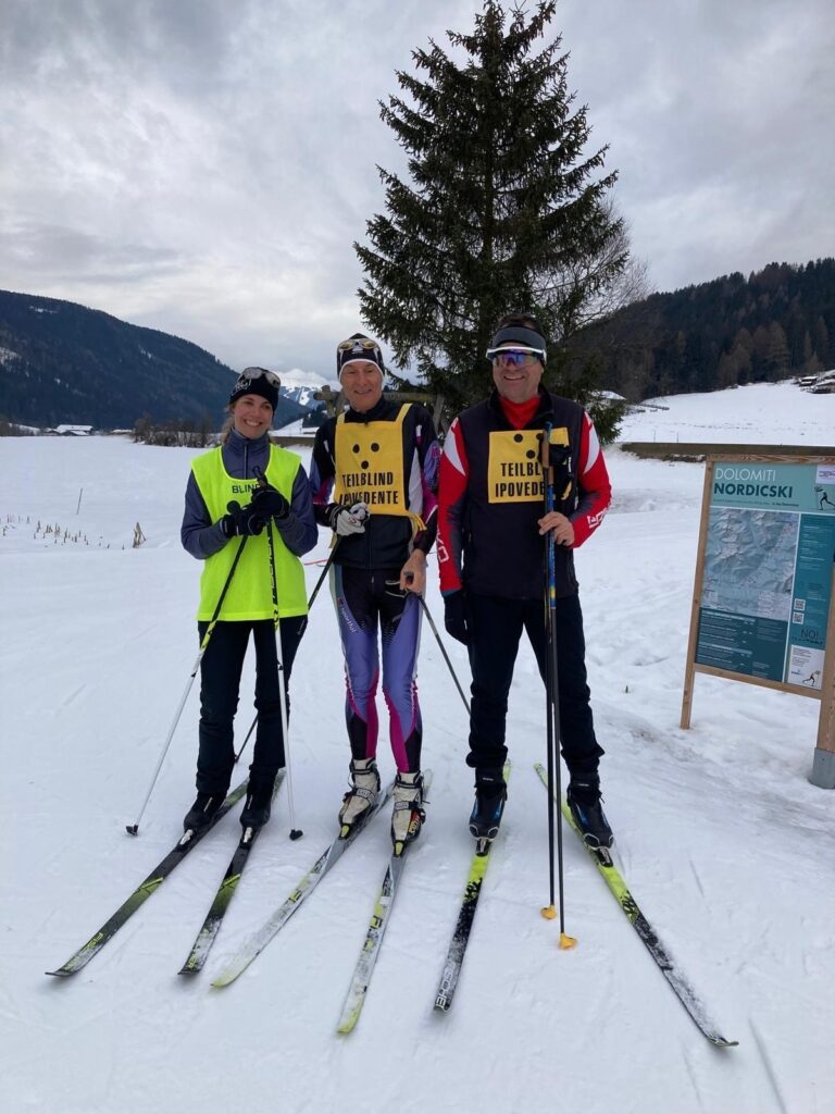 Drei Langläufer stehen lächelnd auf einem verschneiten Feld vor einem Bergpanorama und tragen Westen mit der Aufschrift TEILBLIND IPOVEDENTE.