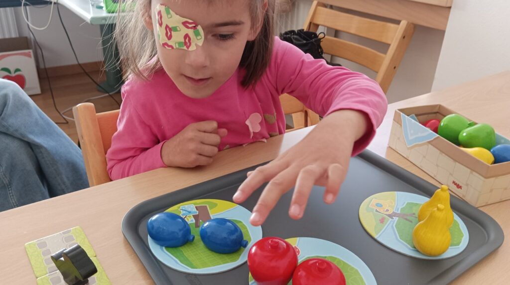 Ein Kind mit Augenpflaster spielt mit buntem Obstspielzeug auf einem Tisch.