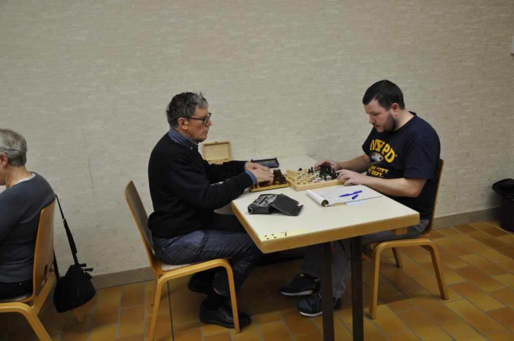 Zwei Männer spielen an einem Tisch Schach, während eine andere Person mit dem Rücken zu ihnen auf einem Stuhl sitzt.