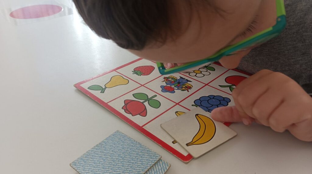 Ein Kind mit Brille spielt mit einem Memory-Spiel, indem es eine Karte mit einem Bananenbild auf ein passendes Bild auf einem Raster legt.