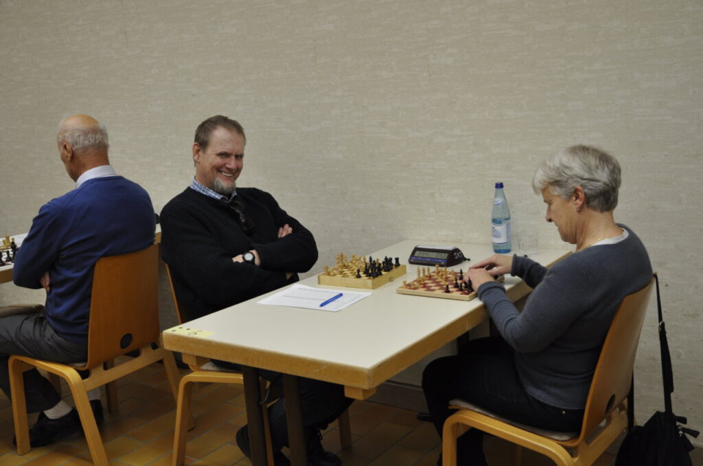 Drei Personen sitzen an Tischen, zwei sind in ein Schachspiel vertieft, während eine Person lächelnd in die Kamera schaut.