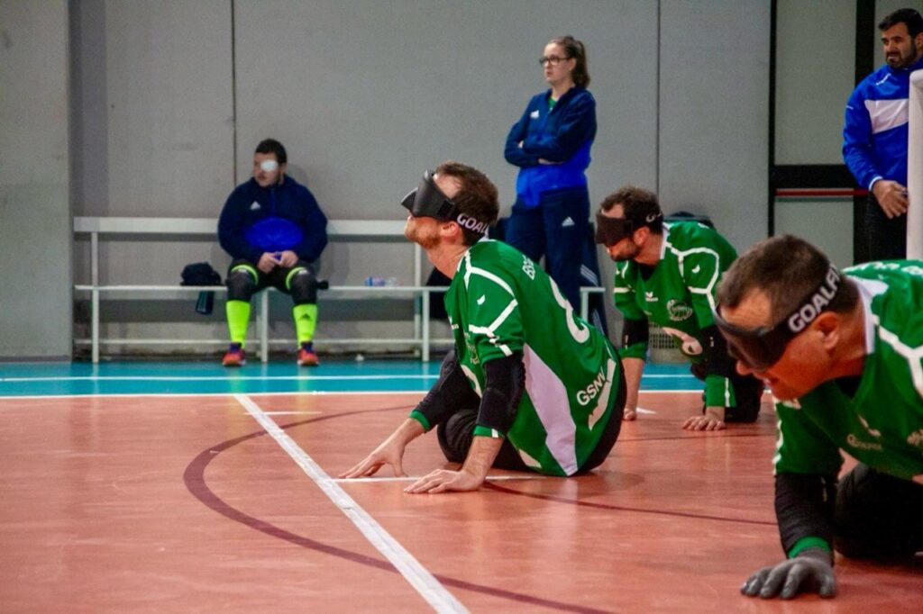 Blinde Sportler in grüner Sportkleidung mit Augenbinden bewegen sich während eines Torball-Spiels auf dem Spielfeld.