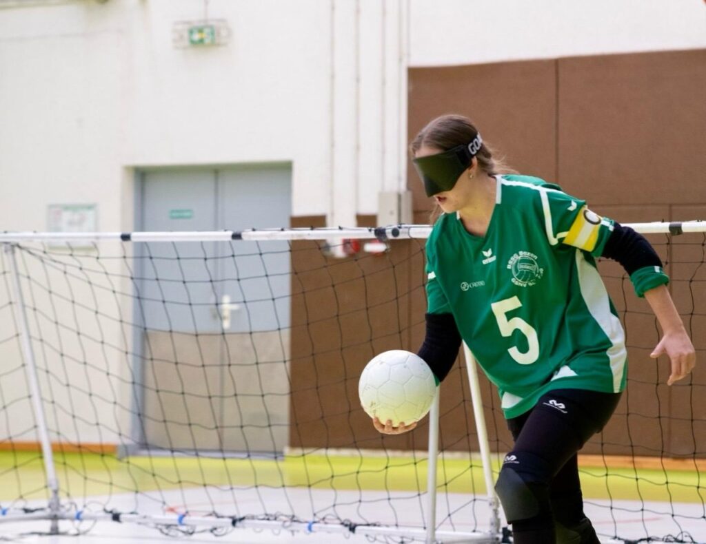 Eine blinde Spielerin in grünem Trikot mit der Nummer 5 hält einen Ball und steht vor einem Torball-Tor in einer Sporthalle.