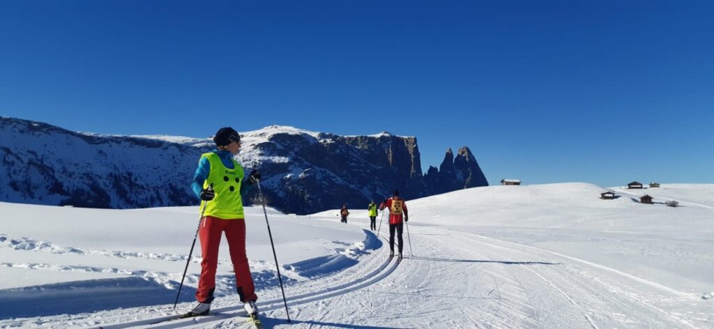 Eine Gruppe von Menschen betreibt bei klarem Wetter Skilanglauf auf schneebedeckten Bergen mit einer markanten Felsformation im Hintergrund.
