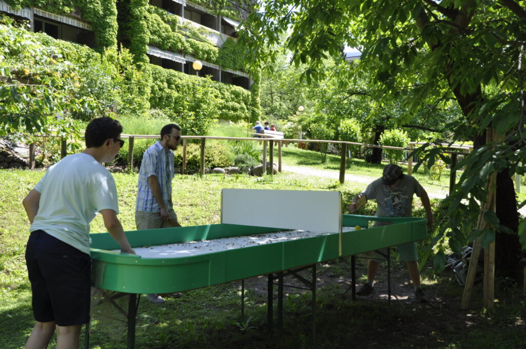 Drei Personen spielen im Freien Tischhockey auf einem grünen Tisch unter Bäumen.