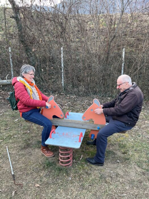 Zwei ältere Erwachsene haben Spaß auf einer Wippe im Freien; ein Taststock liegt auf dem Boden.