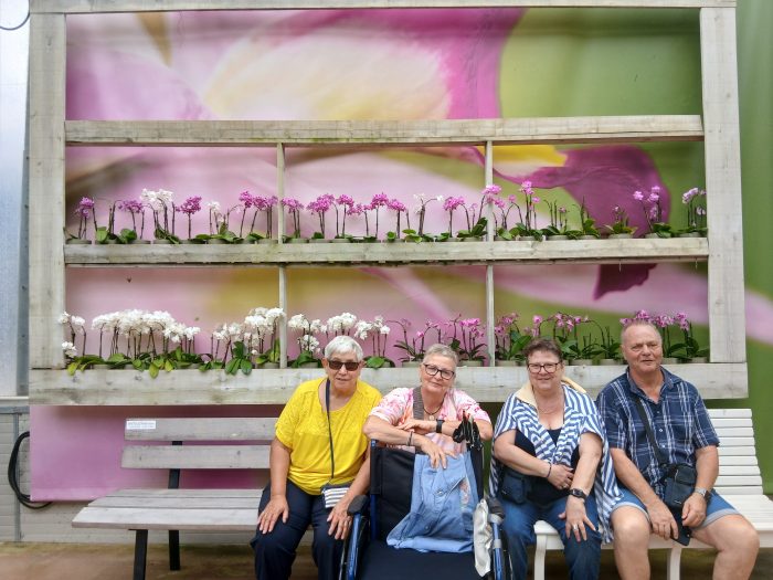 Vier lächelnde Gäste sitzen auf einer Bank vor einer großen Wand mit weißen und rosa Orchideen.