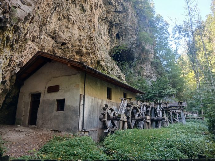 Alte Mühle im Rio-Sass-Canyon unter einer Felswand, davor hölzerne Wasserräder im Grünen.
