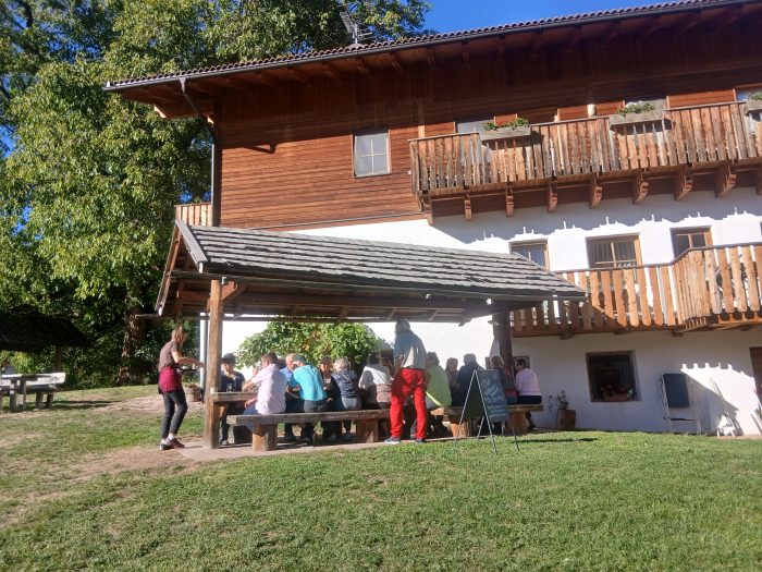 Gruppe von Gästen an langen Holztischen unter einem Vordach vor einem Bauernhaus mit Holzbalkonen.