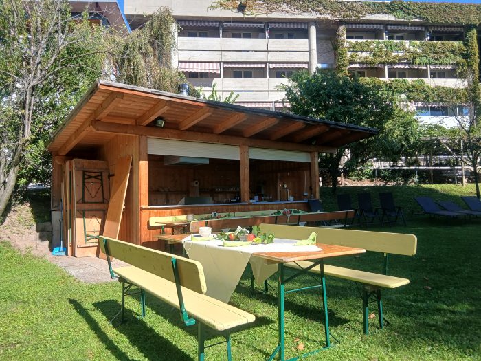 Holzhütte mit gedeckten Tischen und Bänken im Garten, im Hintergrund das efeubewachsene Gebäude des Blindenzentrums.