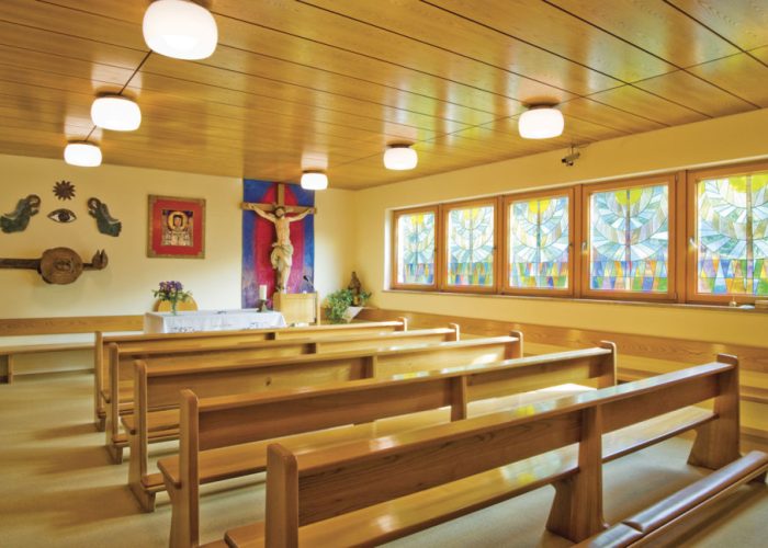 Ein helles, gemütliches Kapelleninterieur mit Holzbänken, bunten Glasfenstern und einem Altar mit einem Kruzifix.