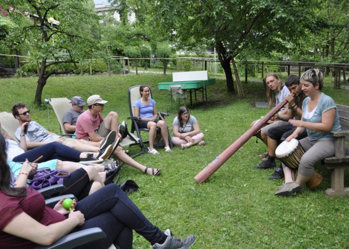 Eine Gruppe von Menschen sitzt in einem Garten im Kreis, während eine Person auf einem Didgeridoo und eine andere auf einer Trommel spielt.