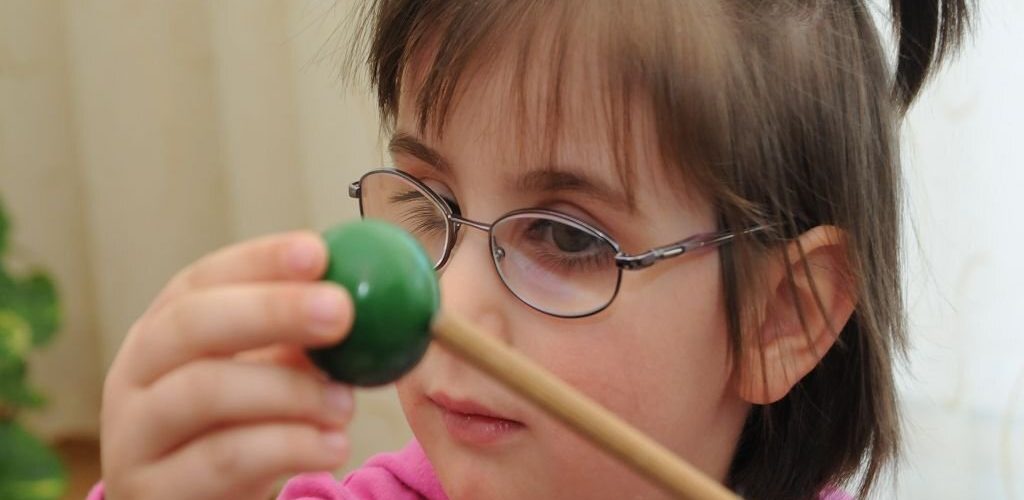 Ein Kind mit Brille hält einen Mallet und betrachtet aufmerksam den daran befestigten grünen Ball.