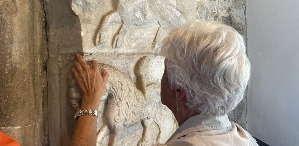 Eine ältere Person mit weißem Haar ertastet mit ihren Händen ein kunstvoll in Stein gemeißeltes Relief.