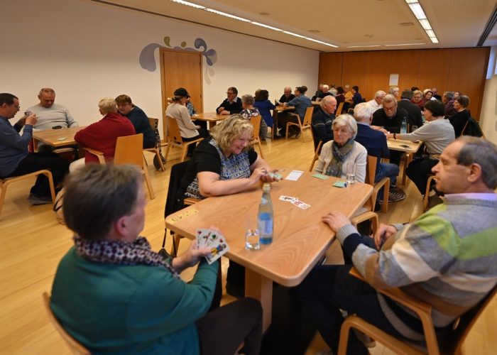 Mehrere Gruppen älterer Menschen sitzen in einem Raum an Tischen und spielen Karten.