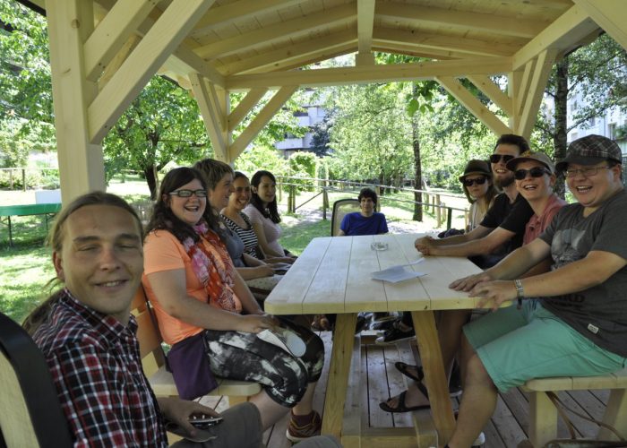 Eine fröhliche Gruppe von Menschen sitzt in einem hölzernen Pavillon an einem Tisch im Freien.