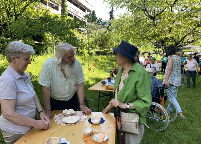 Ältere Menschen stehen und plaudern an einem Tisch im Grünen bei einer Gartenveranstaltung, wobei eine Person Unterstützung durch einen Gehstock hat, im Hintergrund sitzt jemand im Rollstuhl.