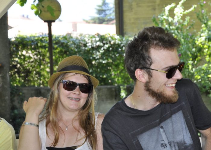 Zwei lächelnde Menschen sitzen draußen im Grünen und tragen Sonnenbrillen.